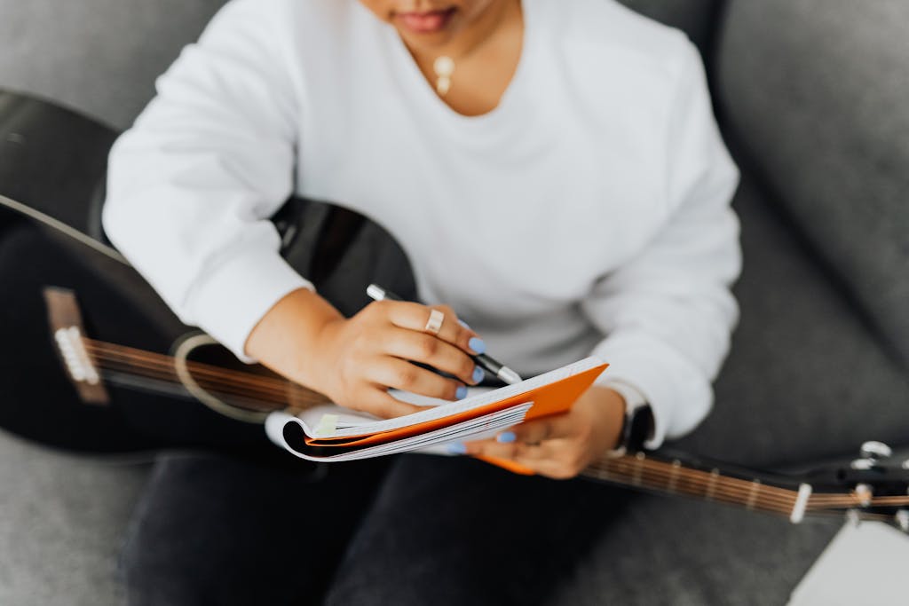 A woman in a white sweater writes music notes in a notebook while holding a black guitar.